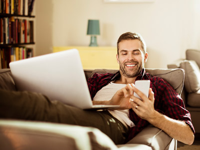 Man laying on his couch checking his account on a mobile phone while is laptop is open on his lap