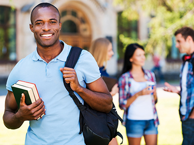 Handsome young African man holding books and smiling while standing against university with his friends chatting in the background